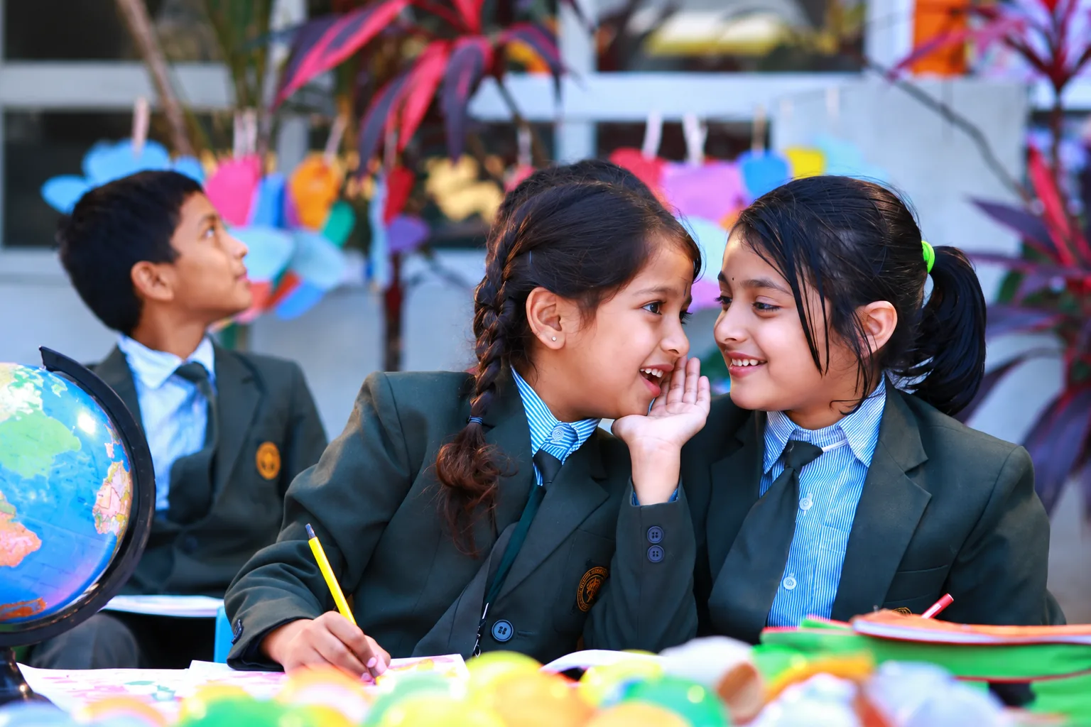 Two Oakwood students whispering together at a classroom desk beside a globe
